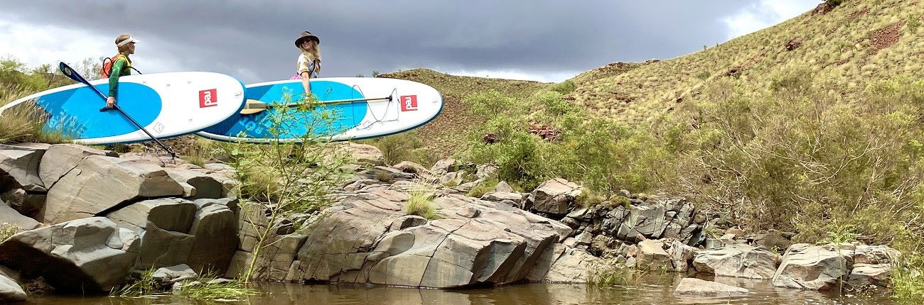 Red Paddle Co SUP lifestyle walking to paddle rocks Stand Up Surf Shop 1300x430